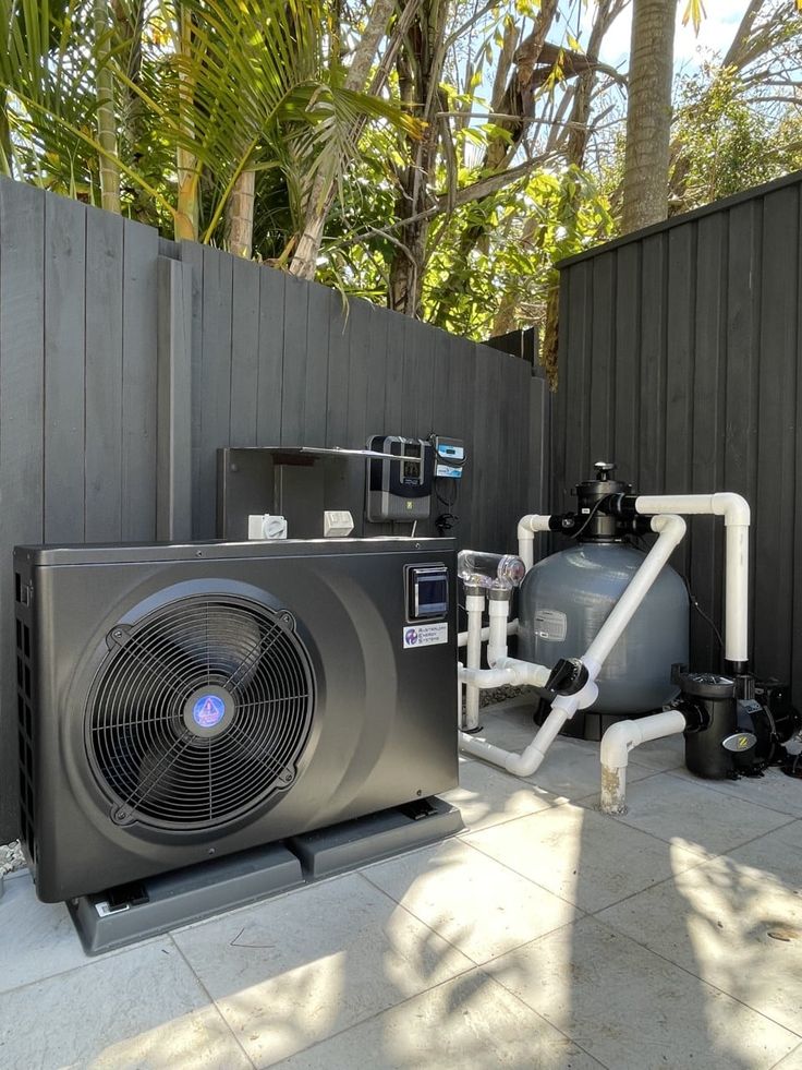 A modern heat pump and pool filtration equipment with white pipes are installed on a tiled outdoor area next to a dark wooden fence, surrounded by tropical plants—an inviting setup for those considering pool heater installation cost. | AES Pool Heating Solutions